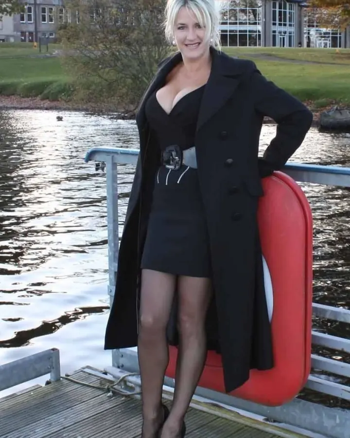Femme aux cheveux gris clair en manteau noir long, robe noire courte et talons aiguilles, souriante, posant sur un ponton près d'un lac.