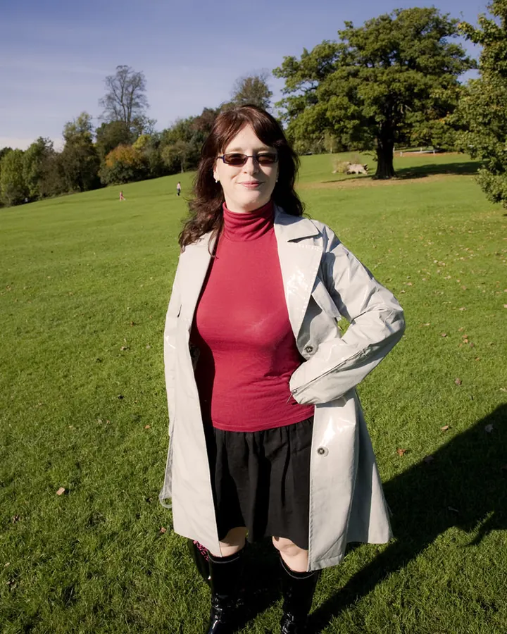 Femme européenne âgée de 45 à 60 ans, portant un pull rouge, une jupe noire, un manteau gris et des bottes noires, posant dans un parc verdoyant.