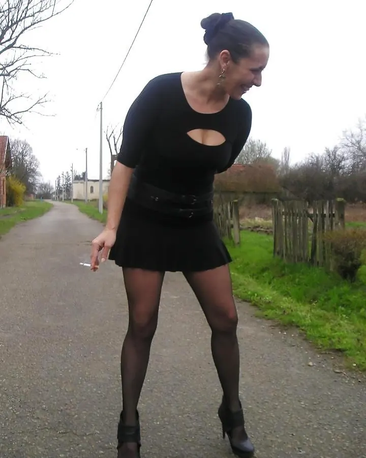 Femme en haut noir et jupe courte noire, marchant sur un chemin rural, tenant une cigarette, portant des collants noirs et des chaussures noires.