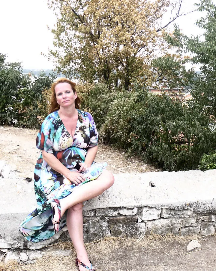 Femme européenne aux cheveux roux, en robe colorée, assise sur un mur de pierre, dans un paysage verdoyant.
