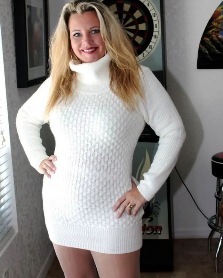 Femme européenne blonde en pull blanc à col roulé, posant les mains sur les hanches, devant un mur avec une cible de fléchettes.