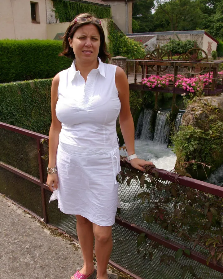 Femme indienne en robe blanche, debout près d'une rambarde, devant une chute d'eau et des fleurs roses, sous un ciel nuageux.