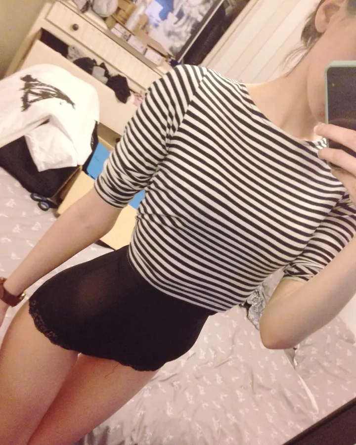 Femme aux cheveux bruns, portant un haut blanc et un short noir, prenant un selfie devant un miroir, dans une chambre.