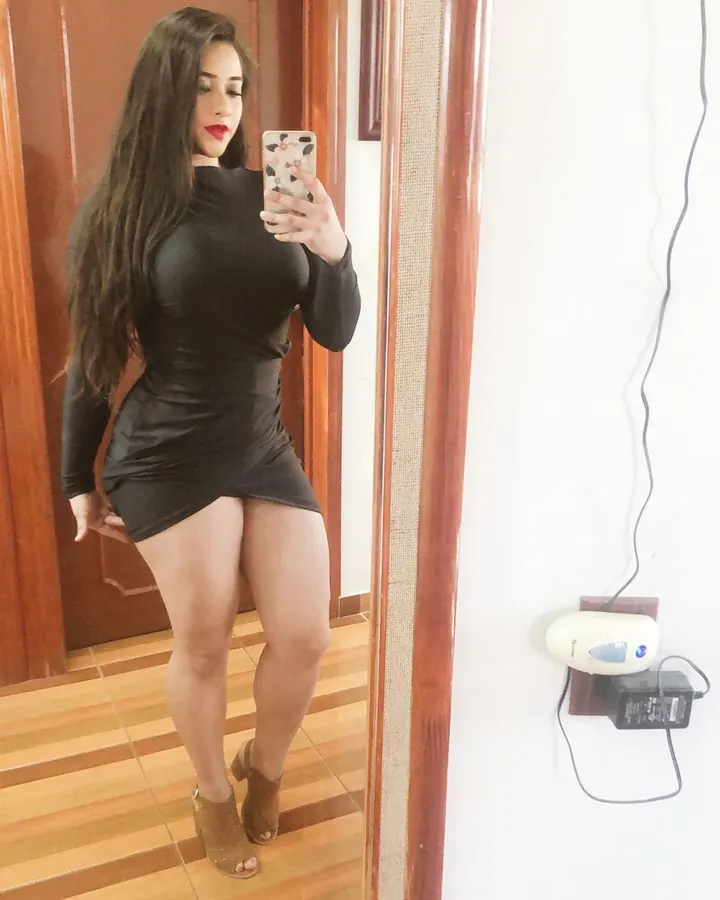 Femme aux cheveux châtains portant un haut noir et un jean déchiré, prenant un selfie dans un miroir avec des bougies rouges autour d’elle.
