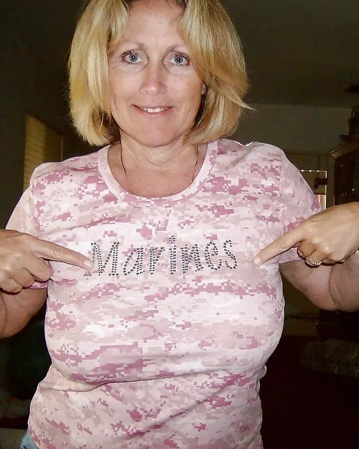 Femme blonde, portant un t-shirt rose camouflage avec le mot "Marines" brodé, pointant du doigt le texte sur son haut.