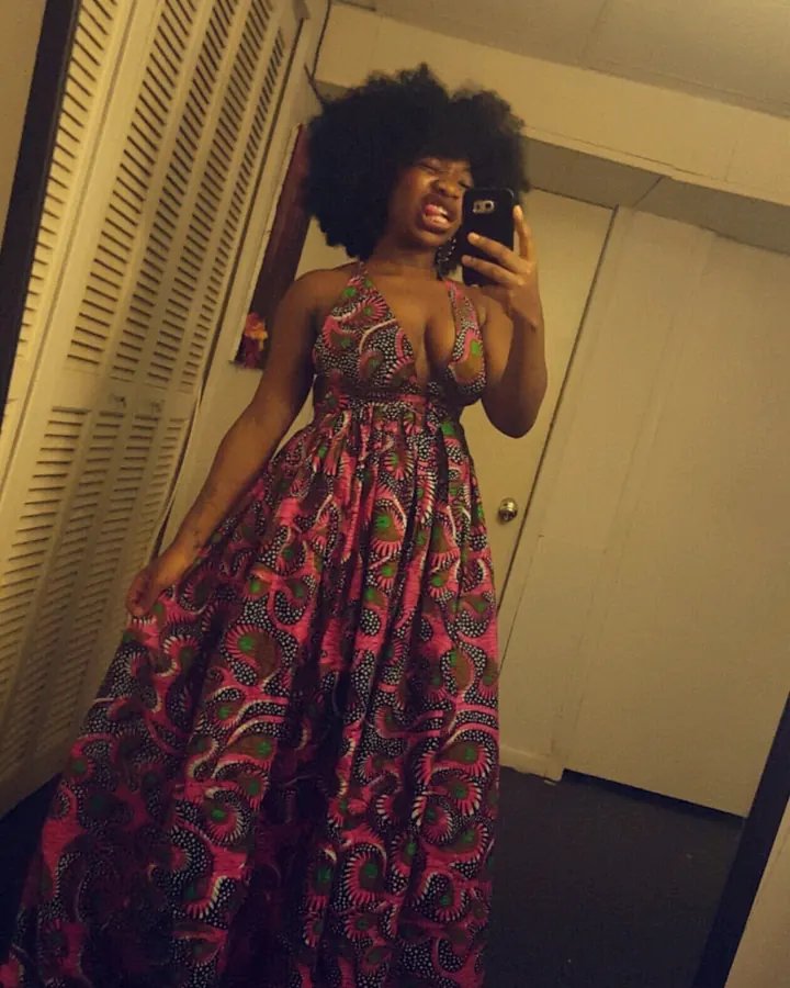 Femme africaine aux cheveux afro, portant une longue robe imprimée rose et verte, prenant un selfie devant un miroir dans une pièce intérieure.