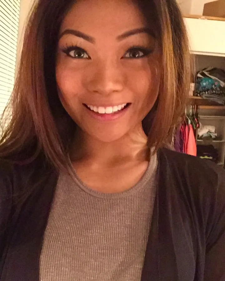 Femme asiatique aux cheveux châtains, souriante, prenant un selfie devant un placard ouvert avec des vêtements à l'intérieur.