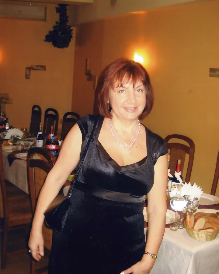Femme en robe noire, assise à une table de restaurant, souriant vers la caméra, main sur la hanche.
