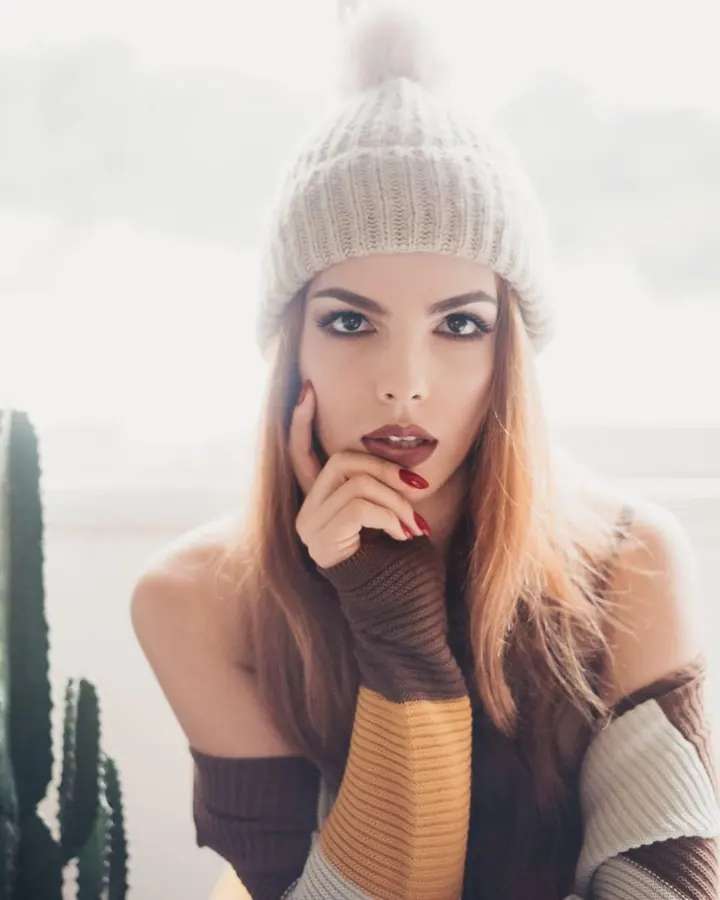 Femme aux cheveux châtains, en bonnet tricoté beige et pull coloré, regardant l'appareil avec intensité, main sur la joue