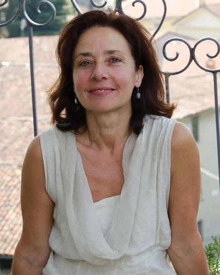Femme aux cheveux châtains, vêtue d’une robe blanche en maille, posant dans une allée de chalets colorés.