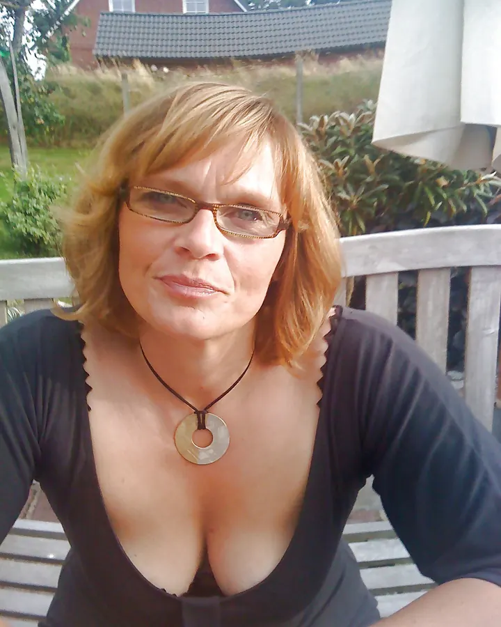 Femme aux cheveux blonds, souriante, vêtue d'une robe noire à col rond, assise sur un banc en bois.