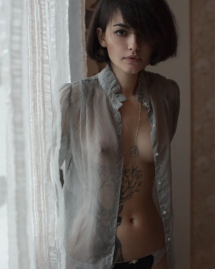 Femme brune en chemise transparente, tatouage arbre, regard vers l’appareil.