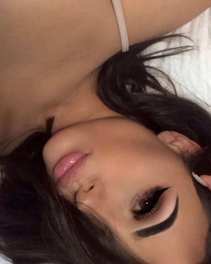 Femme en sous-vêtement noir à dentelle, allongée sur un lit, le visage proche de l'appareil photo, les yeux grands ouverts, une main dans ses cheveux.