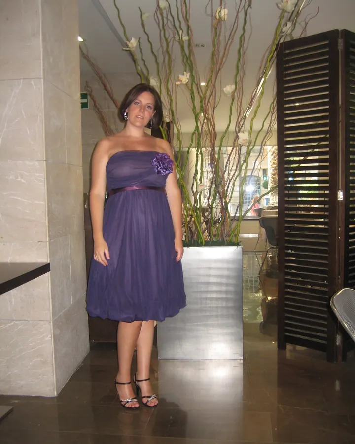 Femme en robe violette sans bretelles, debout dans un hall élégant avec une grande plante décorative derrière elle.