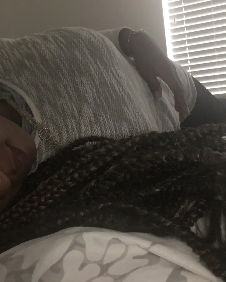 Femme noire allongée, les cheveux en tresses, regardant l'appareil photo, une main posée sur le ventre, dans un lit avec une lumière tamisée venant d'une fenêtre aux volets.