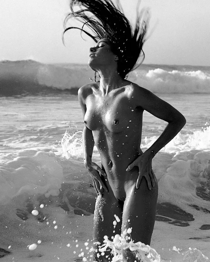 Femme nue sur une plage, cheveux noirs en mouvement, regard vers le ciel, vagues autour d'elle.