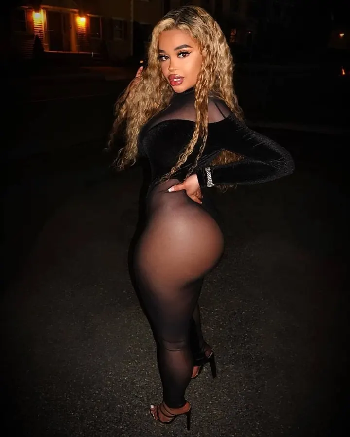 Femme aux cheveux longs bouclés blonds portant une robe noire transparente, debout sur une rue la nuit, les mains sur les hanches.