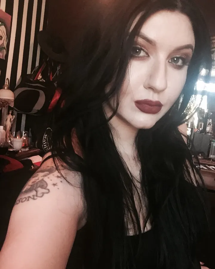 Femme aux cheveux bruns en tenue blanche, posant dans une pièce sombre.