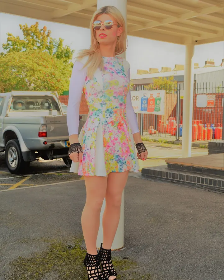 Femme blonde aux cheveux platine, portant une robe fleurie colorée, des lunettes de soleil et des bottines noires, debout sous un abri de station-service.