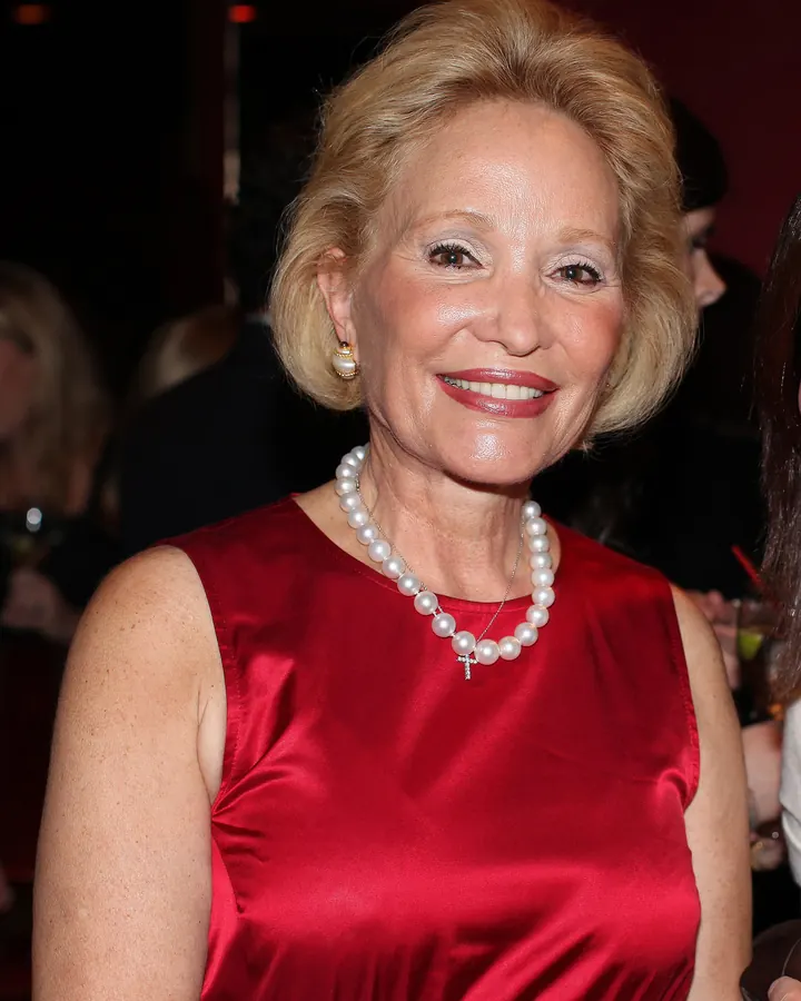 Femme âgée aux cheveux blonds courts, souriante, portant une robe rouge satinée et un collier de perles, dans une ambiance de soirée.