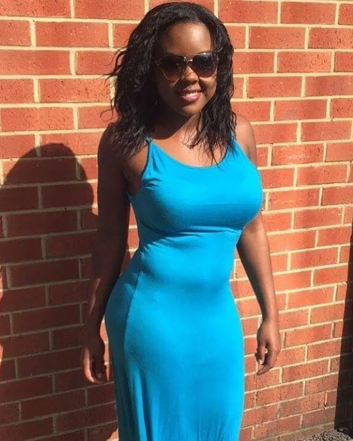 Femme africaine aux cheveux bruns, en robe bleue, posant devant un mur en briques rouges.