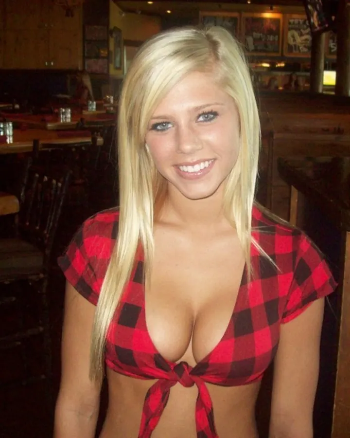 Jeune femme blonde aux cheveux platine, souriante, en tenue de bar, devant un comptoir en bois sombre.