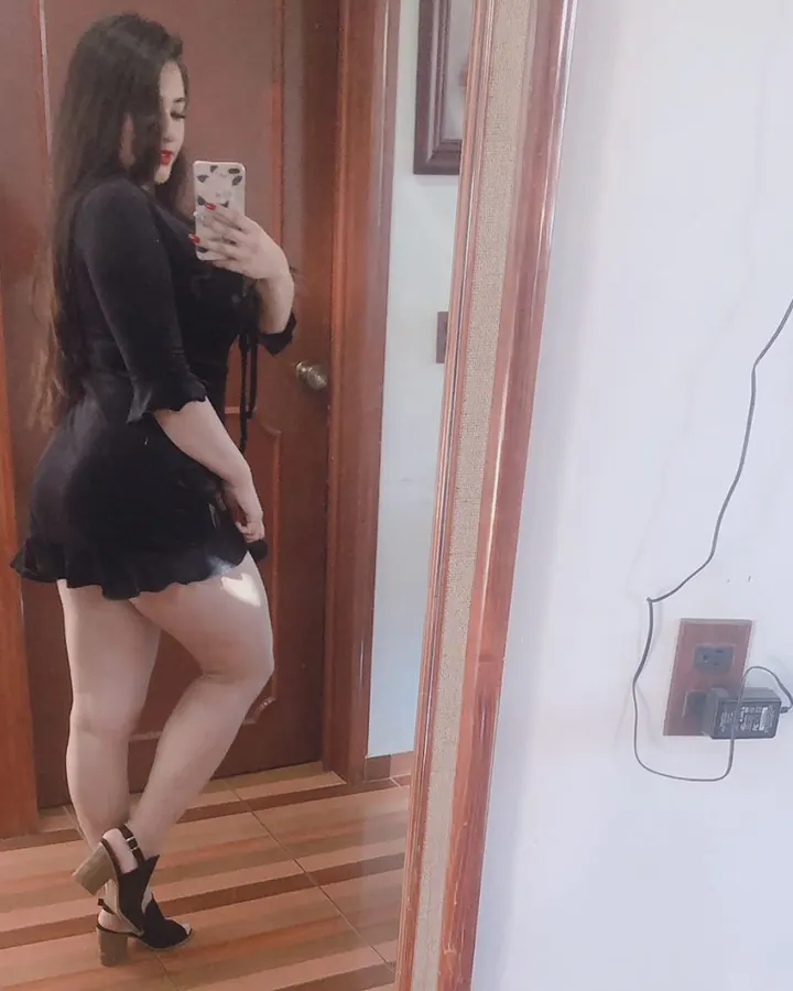 Femme aux cheveux châtains en haut noir, jupe rose, chaussures noires à talons, debout sur un chemin pavé dans un parc.