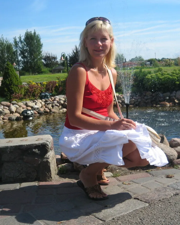 Femme blonde aux cheveux platine, souriant à la caméra, portant un haut noir et une jupe en jean déchiré, prenant un selfie devant un mur blanc.