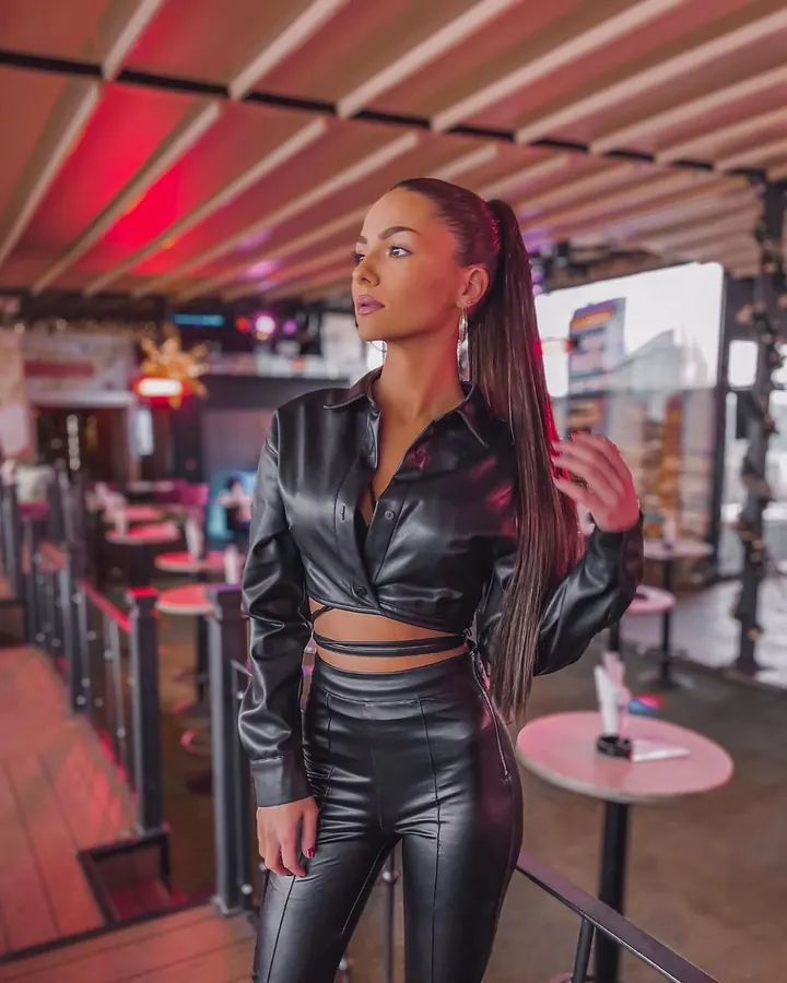 Femme aux cheveux foncés, en tenue en cuir noir, posant dans un bar lumineux, regardant vers le haut