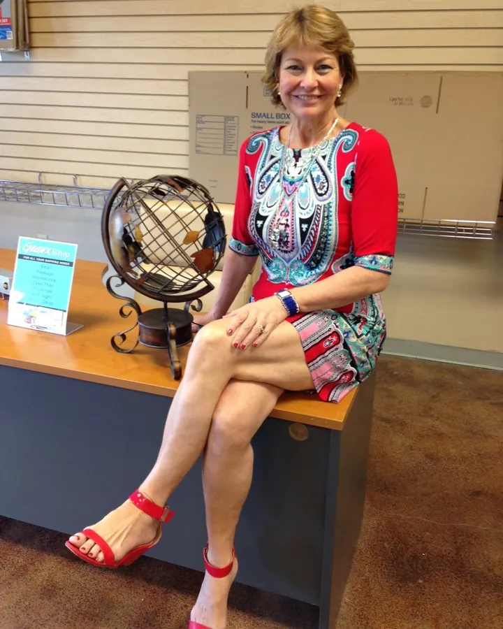 Femme blonde en robe rouge à motifs, assise sur un comptoir, souriante, dans un magasin.