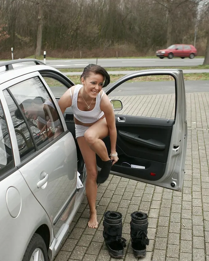 Femme souriante en sous-vêtements blancs sortant d'une voiture, pieds nus, bottes à côté.