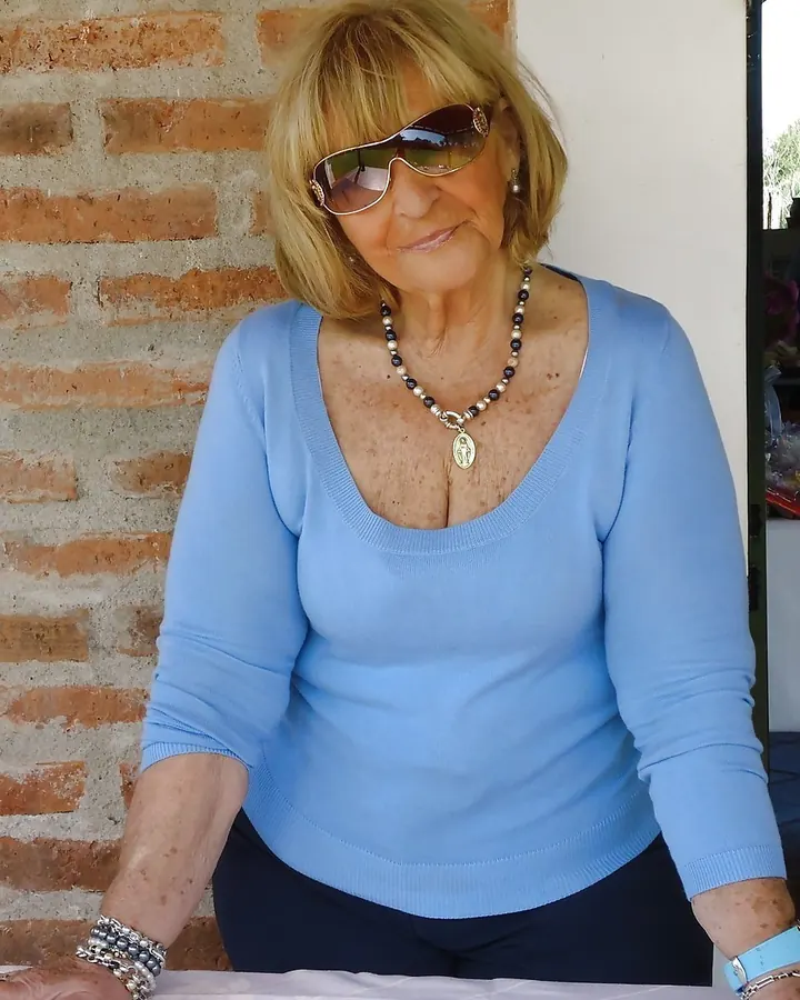 Femme âgée aux cheveux blonds courts, portant des lunettes de soleil, assise contre un mur de briques, vêtue d'un pull bleu clair et d'un collier.