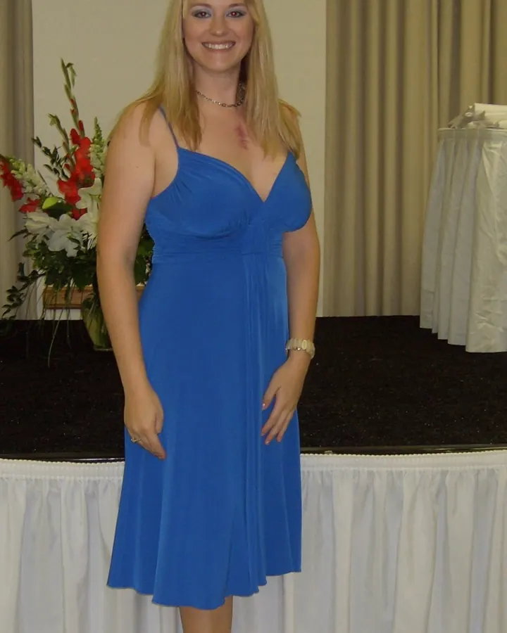 Femme aux cheveux blonds longs, souriante, debout devant une table décorée de fleurs, portant une robe bleue ajustée.