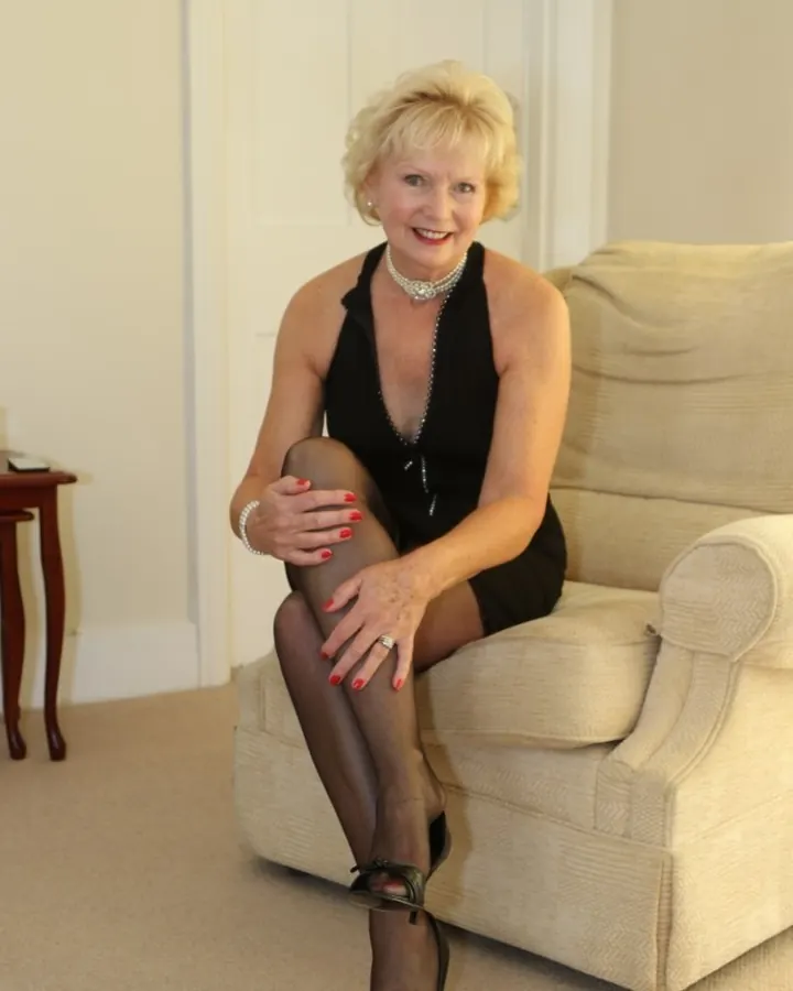 Femme aux cheveux blonds courts, souriante, assise sur un canapé beige, portant une robe noire et des bas noirs.