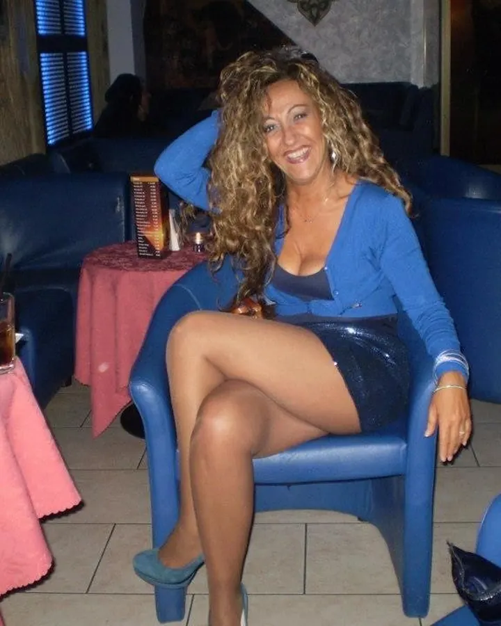 Femme aux cheveux bouclés bruns, souriante, assise sur un fauteuil bleu dans un bar, vêtue d'une veste bleue et d'une jupe courte scintillante.