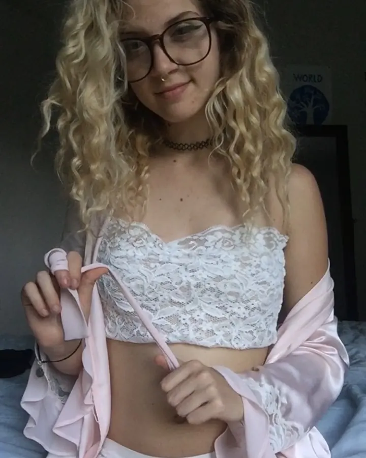 Femme blonde aux cheveux bouclés portant des lunettes, un haut en dentelle blanche et une robe rose pâle, assise sur un lit, tenant un ruban rose.