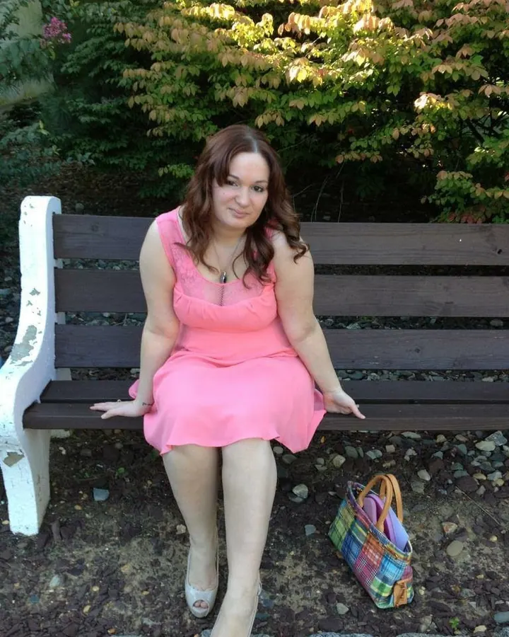 Femme en robe rose, assise sur un banc en bois, dans un jardin, avec un sac coloré à ses pieds.
