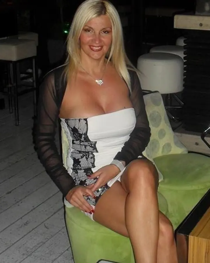 Femme blonde souriante, assise sur un fauteuil vert, vêtue d’une robe blanche à bretelles et d’une veste noire transparente.