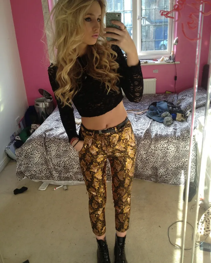 Jeune femme blonde en haut noir ajouré et pantalon doré, prenant un selfie devant un miroir dans une chambre rose