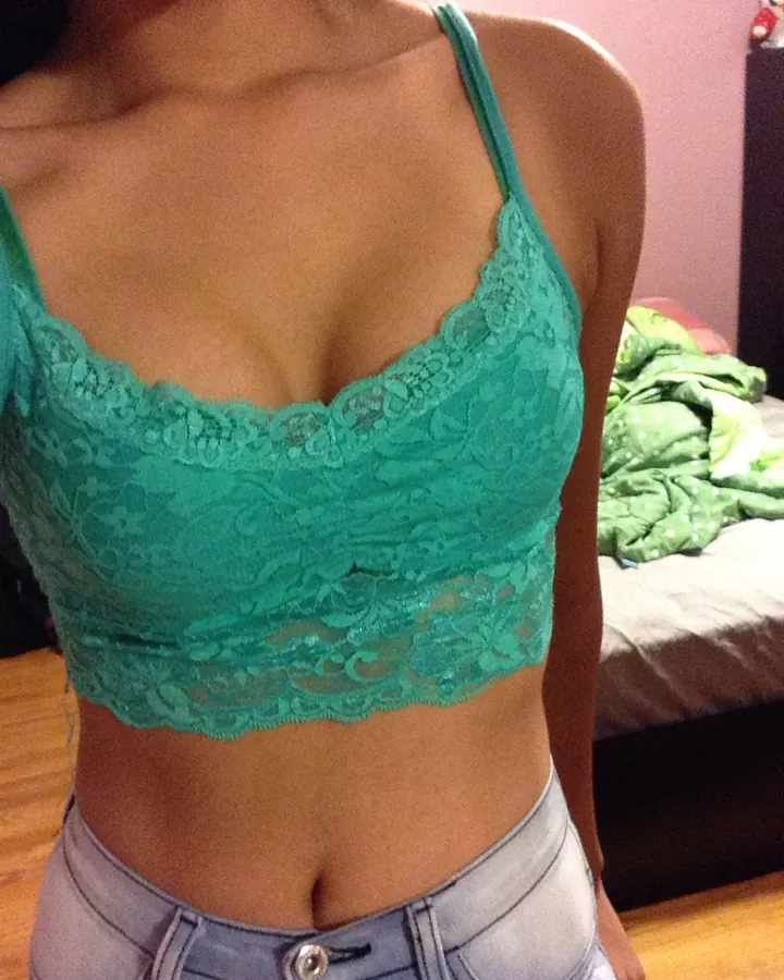 Femme aux cheveux bruns en haut de dentelle turquoise, torse nu, devant un lit.